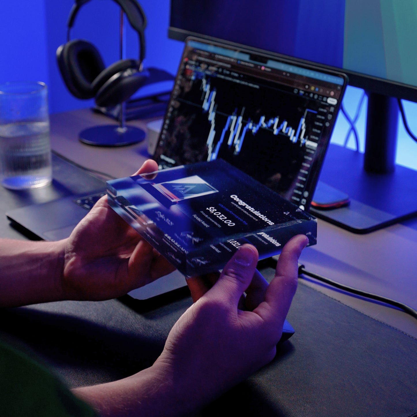 Trader holding a funded award plaque in front of a computer screen with a tradingview charts
