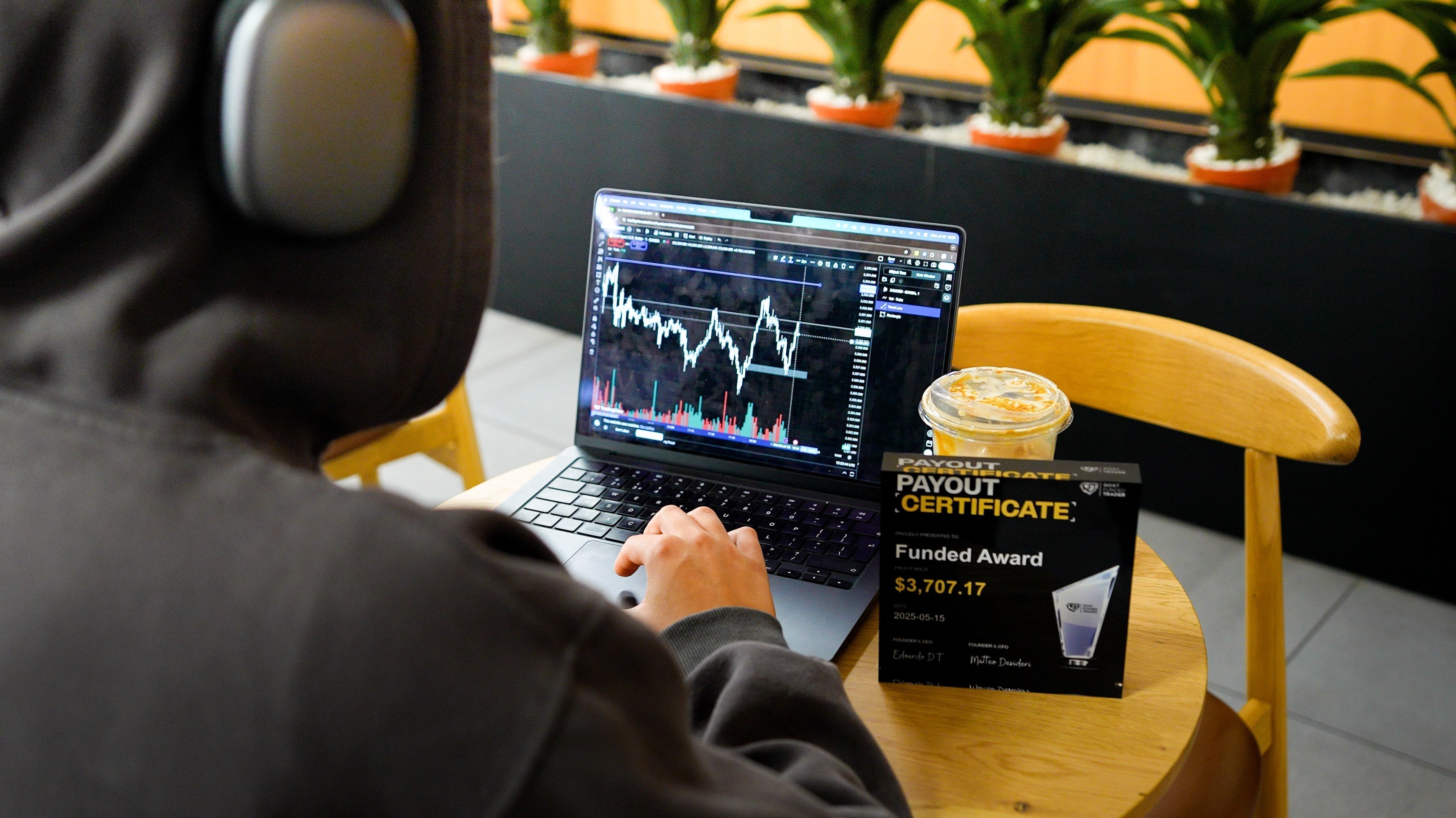 a trader sits in a coffee shop trading on his laptop, having a framed funded certificate to his right hand