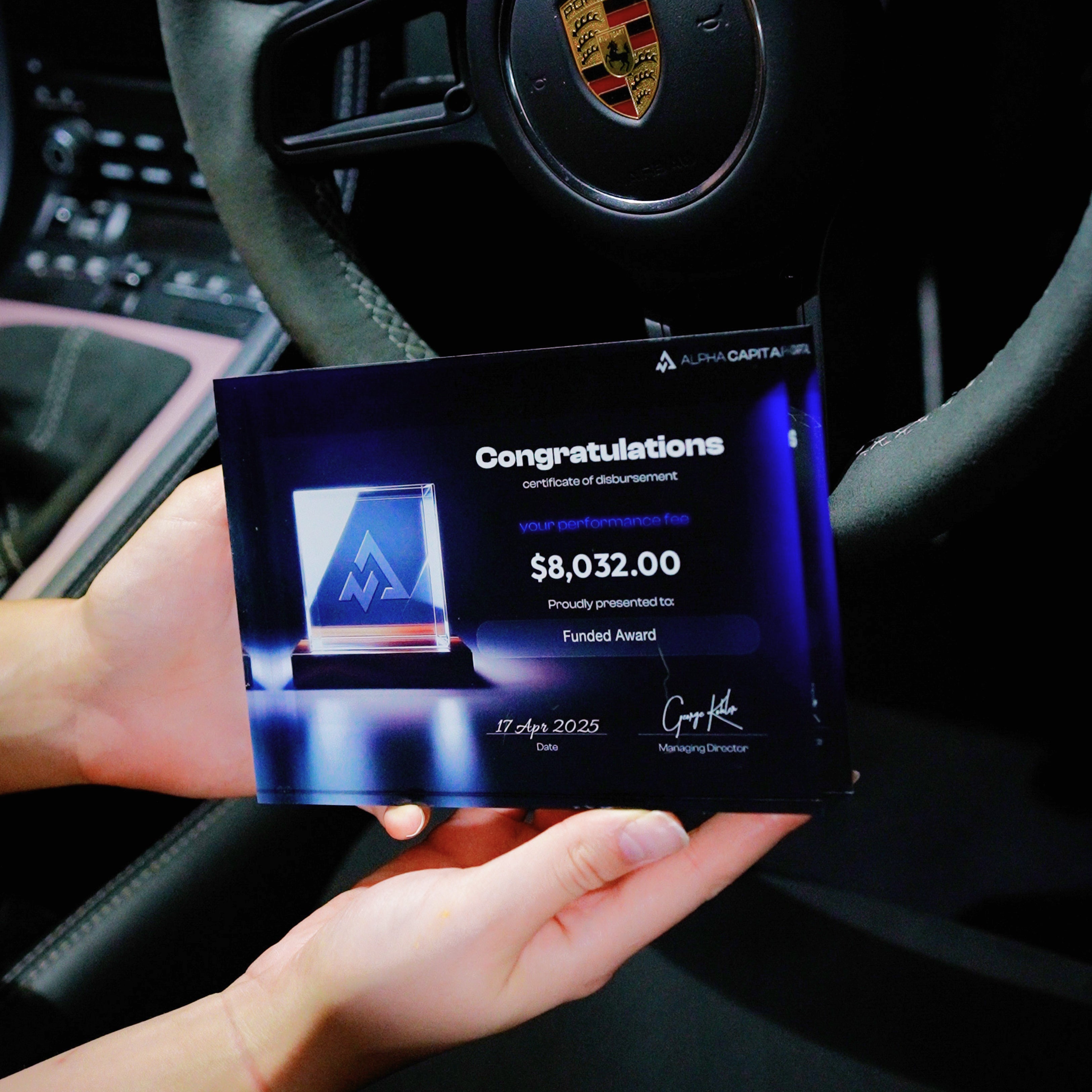 Hand holding apayout Award certificate from Alpha Capital displaying $8,032.00 in a porche car interior.