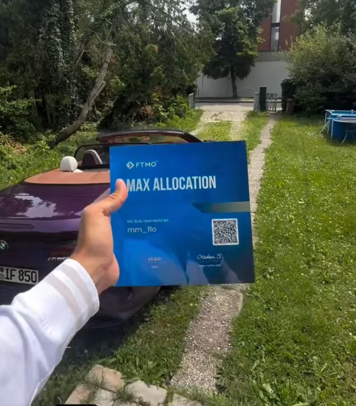 A hand holding a blue Funded Award of FTMO  'Max Allocation' certificate in front of a car outdoor