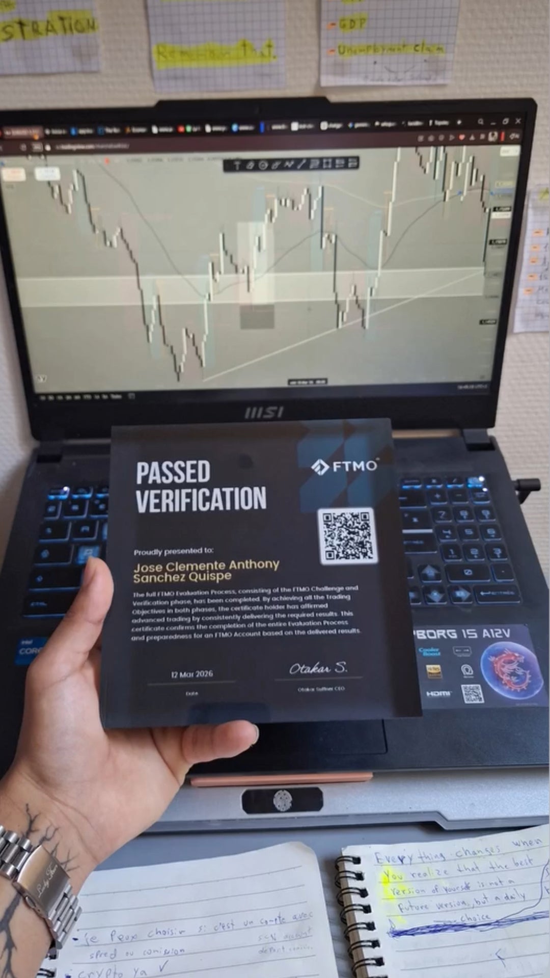 Funded trader holding an FTMO Passed Verification certificate inside a Funded Award plaque in front of a laptop with trading charts