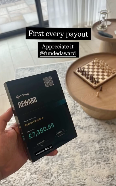 Hand holding an FTMO Reward plaque from Funded Award with a digital display showing £7,350.95, in a room with a chessboard on a table.