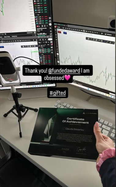 Person holding an Alpha Capital Funded certificate from Funded Award on a desk with multiple computer monitors displaying financial trading data.